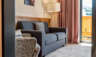 A cozy living room with a gray sofa and a comfortable armchair. Large windows let in plenty of light and provide access to a balcony.