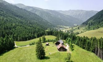 A vast green meadow landscape surrounded by mountains. In the foreground, several cottages stand, surrounded by trees.
