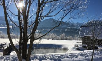 A wintry landscape with snow-covered fields and mountains in the background. The sun shines through the branches of a tree and the water is slightly steaming.