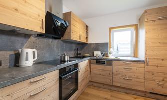 A modern kitchen with wooden furniture and a granite countertop. The room is bright and has a window that lets in daylight.