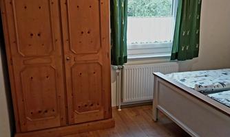 A simple bedroom with a wooden box and a bed. The window has green curtains and lets natural light in.