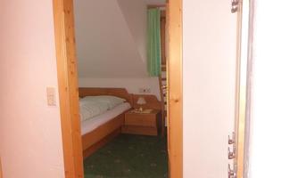 A view into a hallway with an open door frame. In the background, a simple bedroom with a bed and a nightstand is visible.