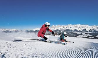 Two skiers are riding down a snow-covered slope. In the background, majestic mountains and a blue sky can be seen.