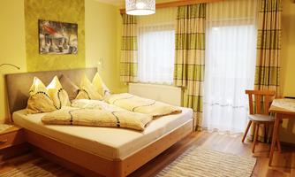A cozy bedroom with a large double bed and yellow pillows. The walls are painted in soft colors, and large windows let in plenty of light.