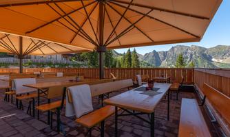 A cozy outdoor area with wooden tables and benches, shaded by large umbrellas. In the background, impressive mountains and a clear blue sky can be seen.