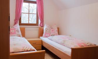 A cozy bedroom with two beds and pink-patterned bedding. Large windows provide plenty of daylight.