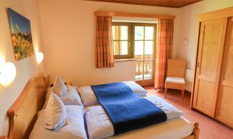 A cozy bedroom with a double bed and a blue blanket. The room has large windows and a comfortable armchair.