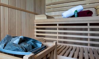 A cozy sauna room with wooden paneling. Towels are lying on a bench, and there is a container with stones.