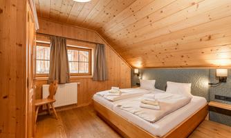 A cozy bedroom with wooden paneling and a double bed. There are windows with curtains and a wooden seating area.