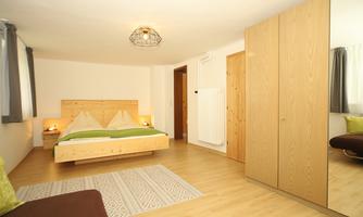 A modern bedroom with a wooden bed and white pillows. The walls are light, and a large wardrobe along with a cozy armchair complement the room.