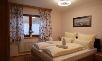 A cozy bedroom with a large bed and soft pillows. In front of the windows are the bright curtains and a small flower arrangement on the windowsill.