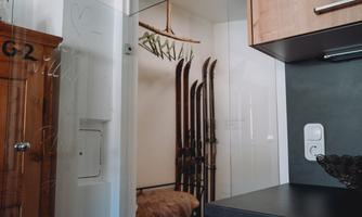 A room with skis on the wall and a wooden cupboard. In the foreground, there is a modern kitchen corner.