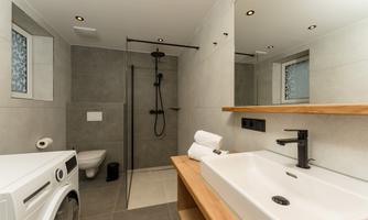 A modern bathroom with gray tiles and a shower. The wooden shelf and washing machine give the room a cozy feel.