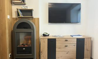 A modernly furnished living space with a TV on the wall and a wooden TV cabinet. Next to the cabinet stands an elegant fireplace for a cozy atmosphere.