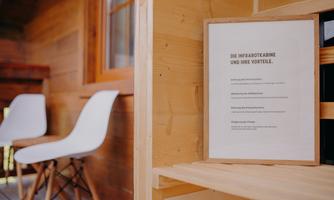 An information board about the advantages of an infrared cabin is attached to a wooden wall. In the background, there are two modern, white chairs.