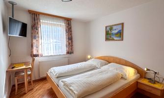 A simple bedroom with two single beds, a desk, and a television. The room has large windows with curtains and a cozy atmosphere.