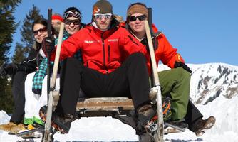 A group of five people is sitting on a sled in the snow. In the background, snow-covered mountains and a clear blue sky can be seen.