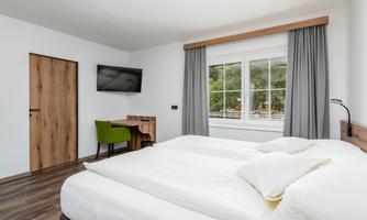 A modern bedroom with two beds, a desk, and a television. Large windows offer a view of the greenery.