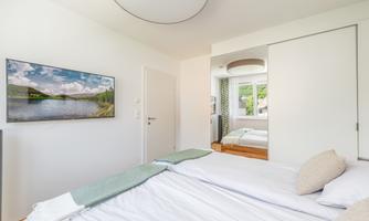 A bright bedroom with two beds and simple modern design. A picture of a landscape hangs on the wall and a window offers a view of nature.