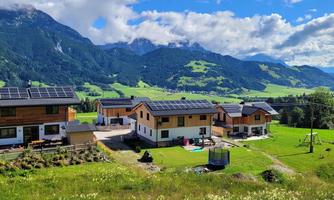 A picturesque landscape with modern houses and solar panels. In the background, green meadows and majestic mountains stretch under a blue sky.