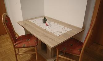 A small dining area with a wood-colored table and two chairs. On the table is a decorative tablecloth with a small plant.