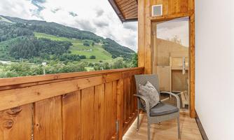A cozy balcony with wooden paneling and a chair. In the background, a green mountain landscape stretches out.