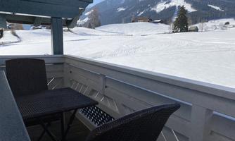 A wooden terrace with two chairs and a table. In the background, snow-covered hills and a blue sky are visible.
