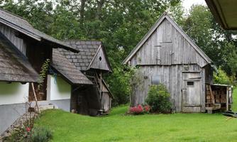 A quiet, rural farm with old wooden houses and fresh, green grass. Surrounded by trees and blooming plants, the landscape appears inviting and idyllic.