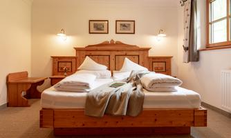 A cozy bedroom with a wooden bed and two pillows. The walls are painted in light colors, and there is a desk as well as a large window with natural light.