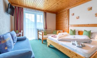 A cozy room with wooden paneling, a large bed, and a sofa bed. There are large windows with daylight and colorful cushions on the beds.