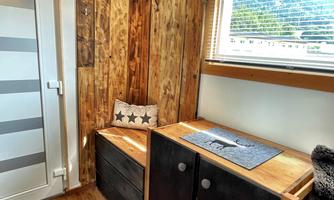 A cozy room with wooden walls and a windowsill that lets in plenty of light. There is a seating area with wooden shelves and neatly arranged items.