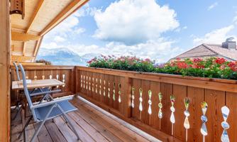 A beautiful balcony with a wooden railing and flowers. In the background, there are mountains and a blue sky with clouds.