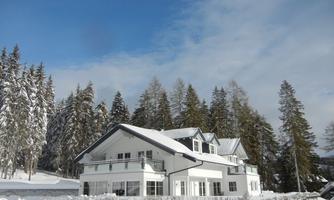 A modern house in a snowy landscape surrounded by tall trees. The sky is clear with a few clouds.