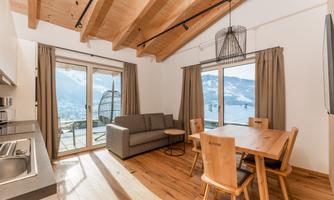 A modern living room with a wooden beam ceiling, a gray sofa, and a dining table. Large windows offer a view of the mountains.