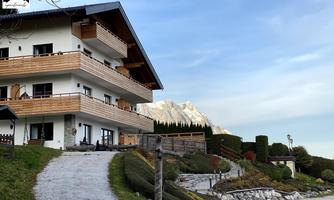 A modern house with a balcony and wooden cladding, surrounded by well-maintained gardens. Impressive mountains can be seen in the background.