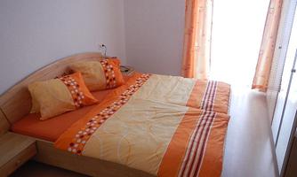 A cozy bedroom with a double bed and orange bedding. The large window front lets in plenty of daylight.