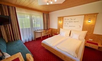 A cozy hotel room with a double bed and wooden furniture. There is a table, a television, and large windows that let in daylight.