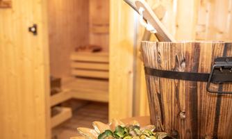 A rustic wooden container stands in the foreground, while a sauna is visible in the background. The atmosphere feels relaxing and inviting.