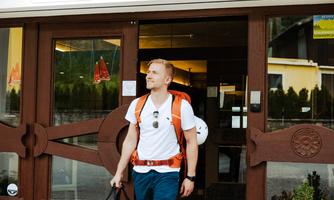 A traveler leaves the Mountain Hostel with a backpack and a bag. In front of the hostel, colorful flowers are in a pot.