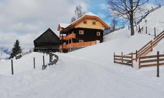 A snow-covered landscape with two small cottages. In the background, there are trees and a cloudy sky.