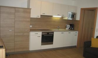 A modern kitchenette with white cabinets and wooden details. The kitchen is equipped with a stove, a microwave, and a coffee machine.