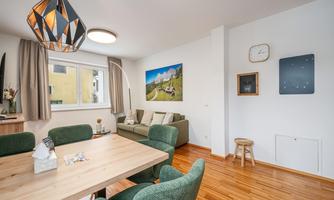 A modern living room with a wooden table and green chairs. Pictures hang on the wall, and a lamp provides pleasant light.