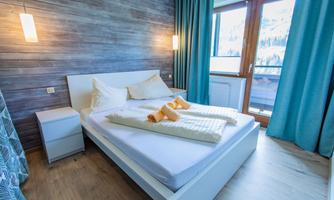 A modern bedroom with a large bed and bright bed linens. Large windows let in plenty of light and offer a view outside.
