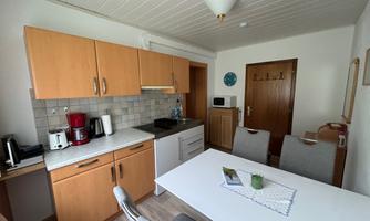 A modern kitchen with wooden cabinets and a white dining table. There are cooking appliances, a coffee machine, and a cozy entryway.