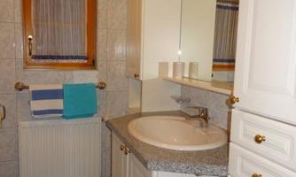 A modern bathroom with a sink, a mirror, and simple white cabinets. The walls are tiled, and there are colored towels and rugs.