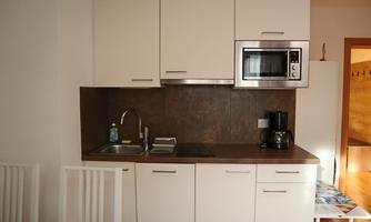 A modern kitchen unit with white cabinets and a dark countertop. It is equipped with a sink, microwave, and coffee machine.
