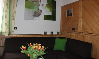 A cozy living room with a dark couch and a vase full of yellow tulips. A modern painting hangs on the wall, and there are wood paneling and large windows with green curtains.