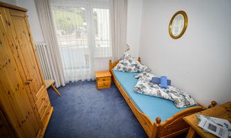 A cozy room with a single bed, a wooden wardrobe, and a table. The large windows let in plenty of light.