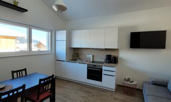 A modern kitchen with a dining table and chairs. Next to the kitchen is a cozy sofa, and large windows bring a lot of light into the room.