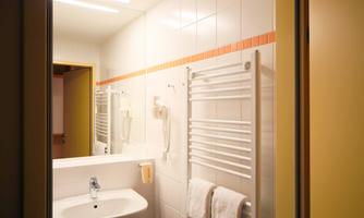 A modern bathroom with white tiles and a sink. Towels hang on a towel rack, and there is a large mirror.